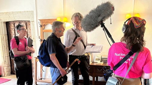Two podcast producers with clipboards and boom mikes record the presenter and speaker in a yellow room